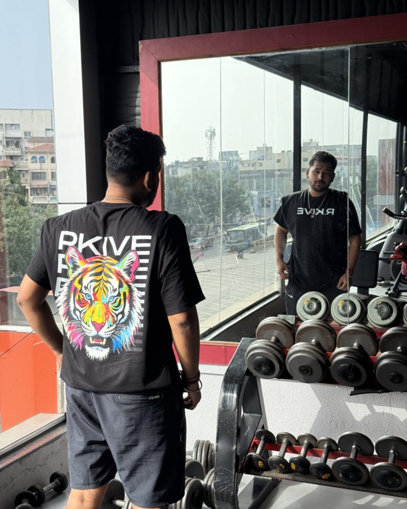 A gym-goer wearing a bold black graphic T-shirt featuring a vibrant multicolor tiger design and "RKIVE" branding, reflected in the mirror inside a modern fitness studio.
