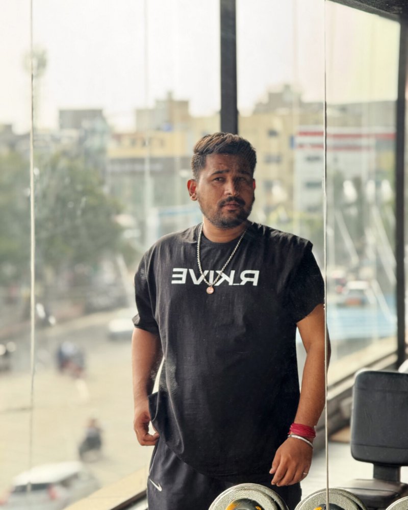 A man wearing a black Swavable T-shirt with “RKIVE” text, standing confidently near a gym mirror with city buildings and traffic visible through large glass windows.